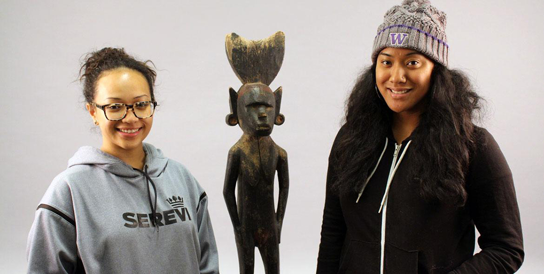 Two young woman stand beside a small wooden statue of a person