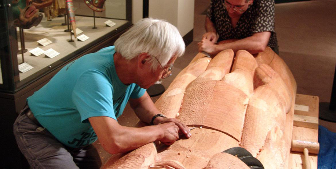 A man carves a house post out of wood inside of a museum gallery
