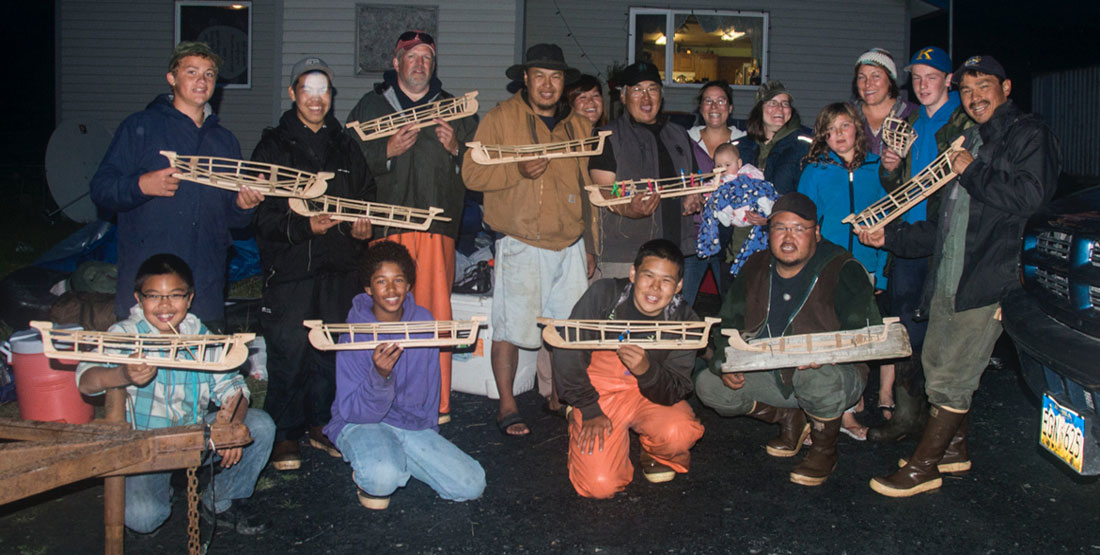 A large group of people stand outside holding model Angyaaq boats