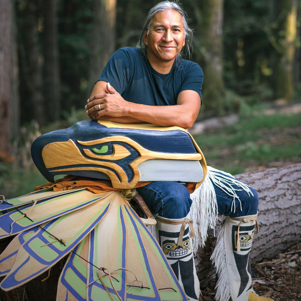 A man in a blue shirt sits with a mask that looks like the Seahawks logo and ceremonial clothes that look like wings