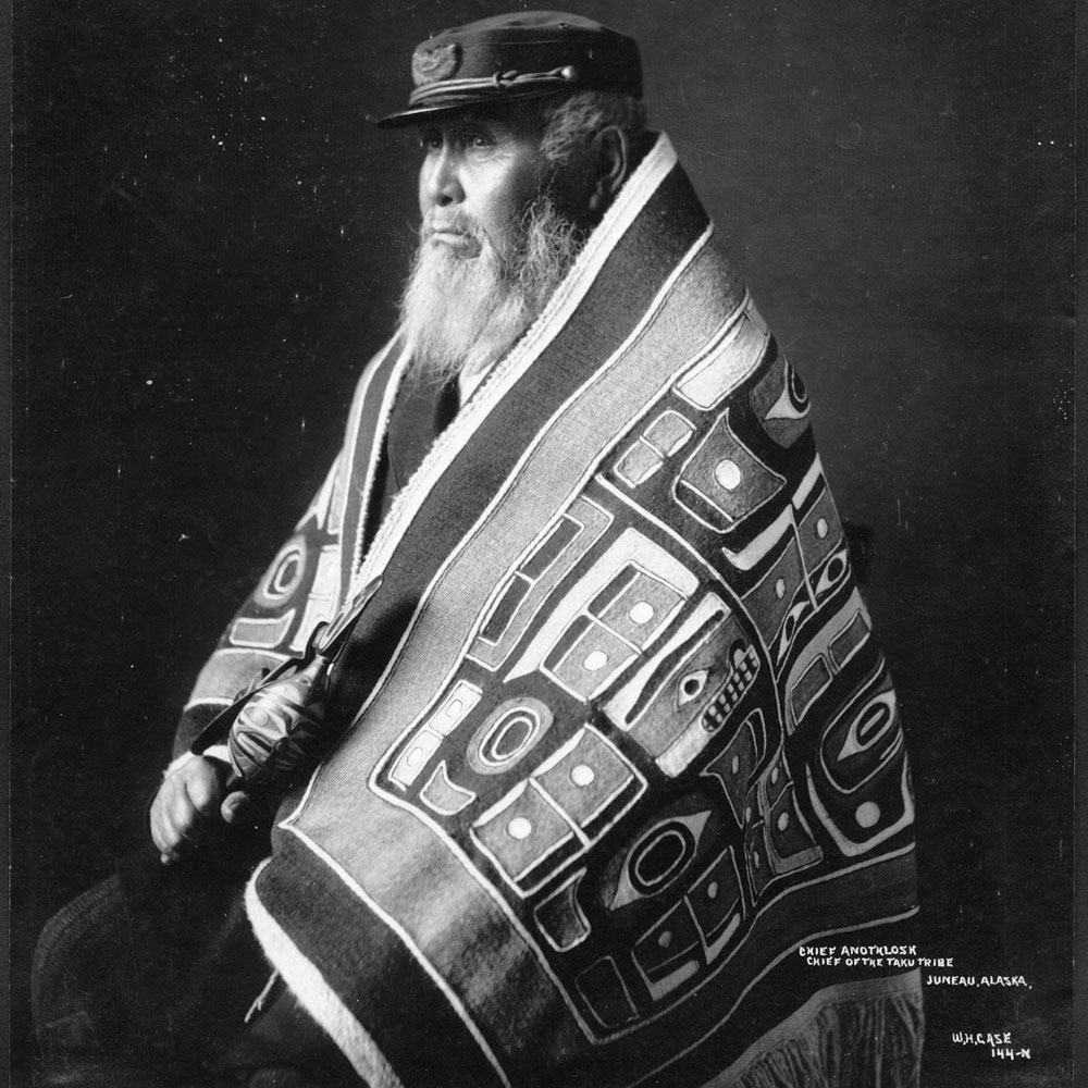 An historic black and white photograph of an old man wrapped in a decorative and fringed blanket