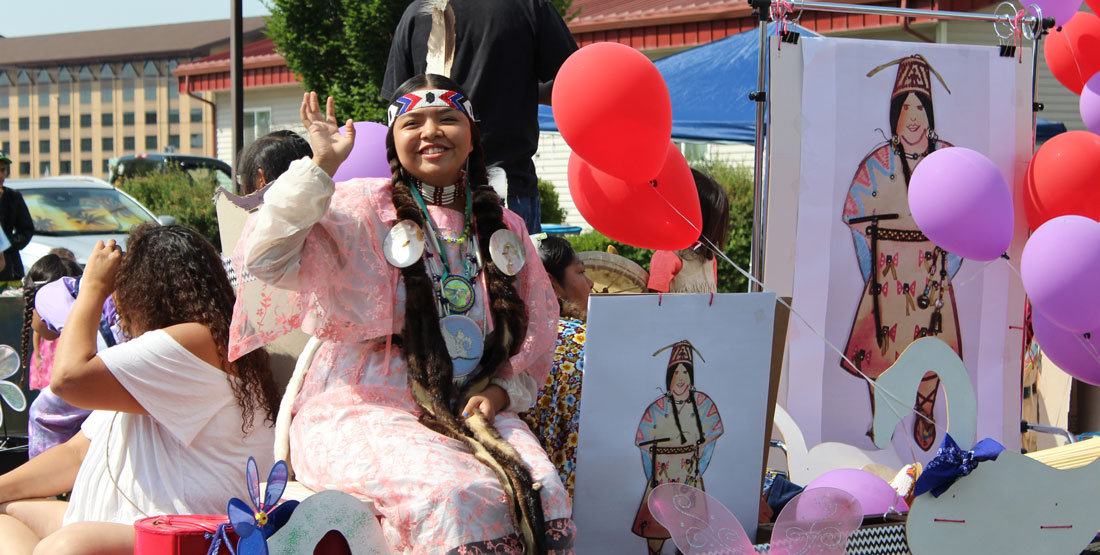a young woman wearing traditional regalia smiles and waves from a float in a parade