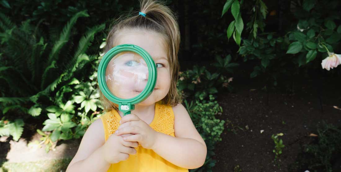 a kid holds a large green magnifying class up to her eye, making her eye appear much larger