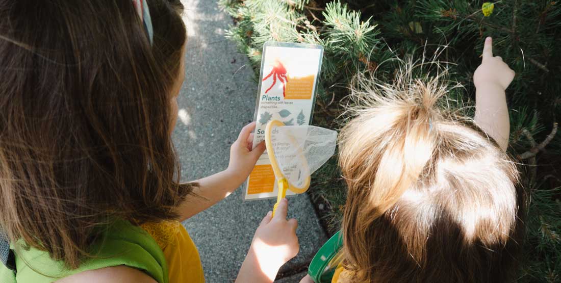 two kids engage with the nature scavenger hunt. one points to a leaf on the paper, one points to it on a tree
