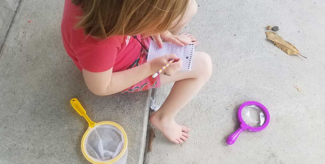 a kids writes in a tiny notebook while using the materials in a backyard scientist backpack