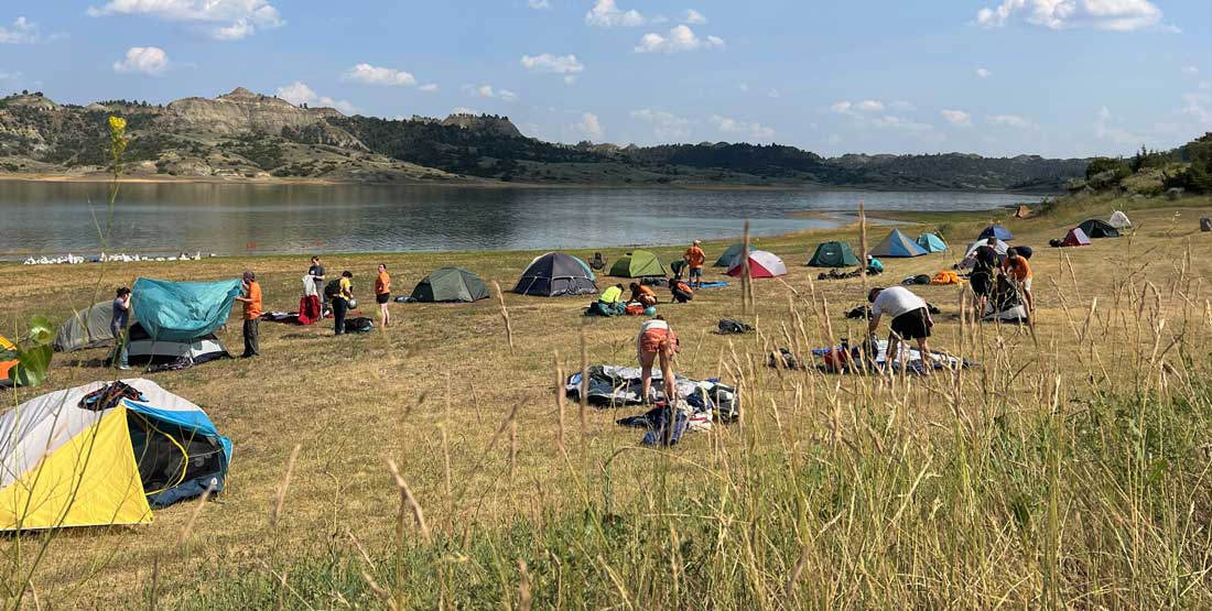 The grassy shore of a lake is dotted by several multi-colored camping tents. Mountains and blue sky can be seen in the background