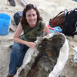 holly woodward kneels next to a fossil in a plaster jacket