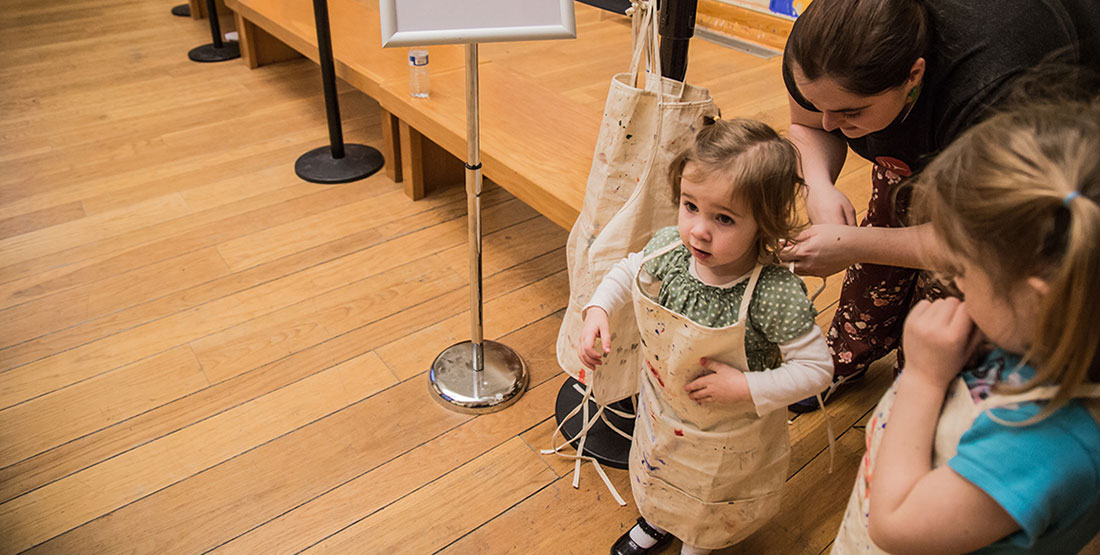 A young toddler girl waits patiently as she has an apron tied around her waist