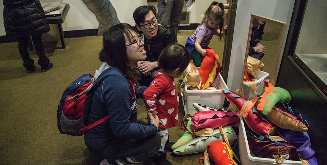 A family prepares to put a dino tail on their toddler in the dino dress up corner
