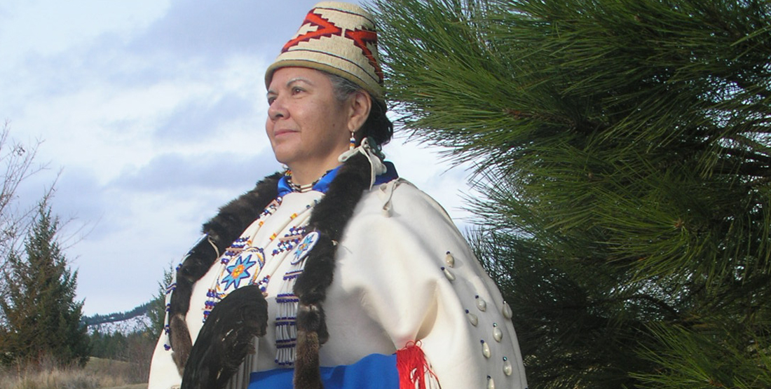 A woman stands by a tree wearing full regalia