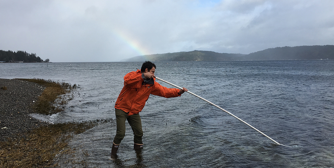 A man stands on the beach dipping a pole into the water. A rainbow is behind him.