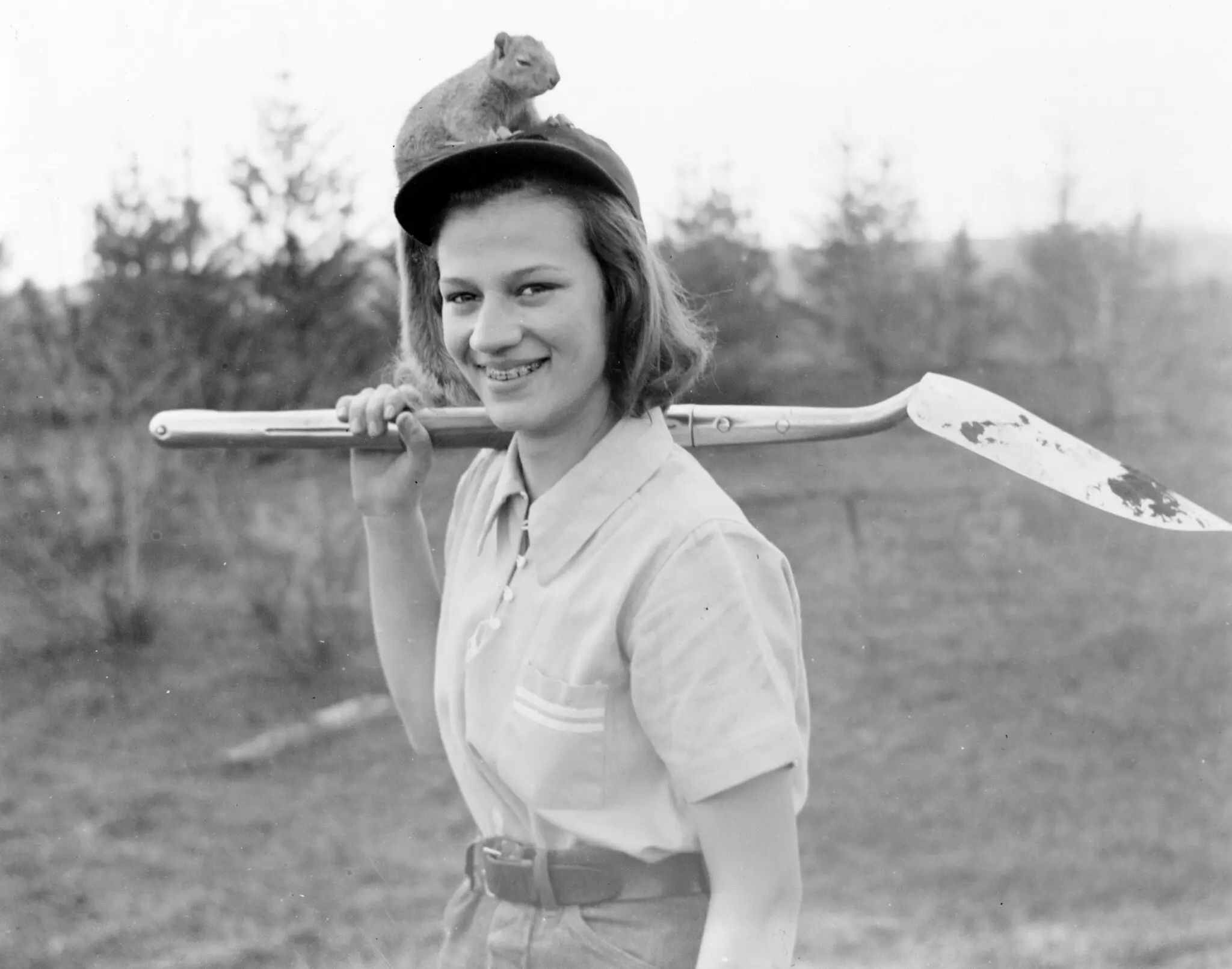 Woman in a black and white photo with a shovel over her shoulder smiles at the camera. A squirrel sits on top of her head.