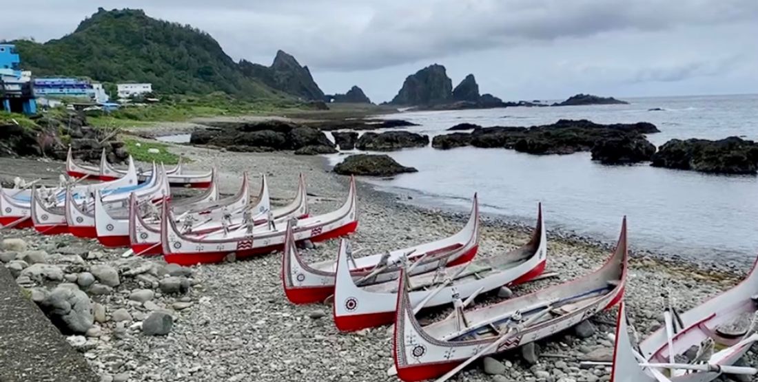 Brightly painted boats lined up on a beach with the ocean and grassy rocks in the background.