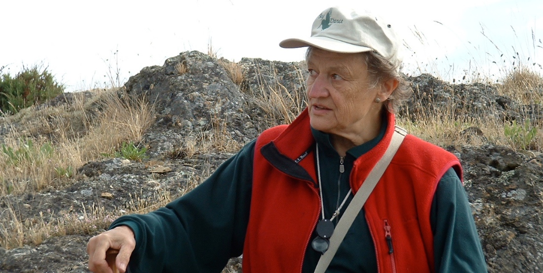 Woman in a red vest and green shirt with rocky grasslands in the background. She wears a hat and looks toward the left side of the frame.