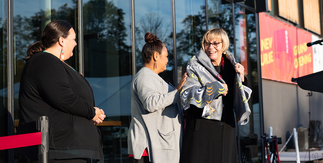 three women stand in front of the new burke museum building, one is wearing a blanket that says "burke" on it