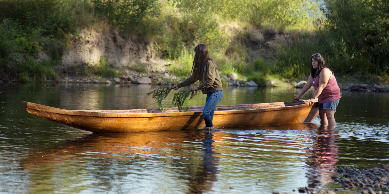 two women place cedar branches in a carved canoe floating on the water