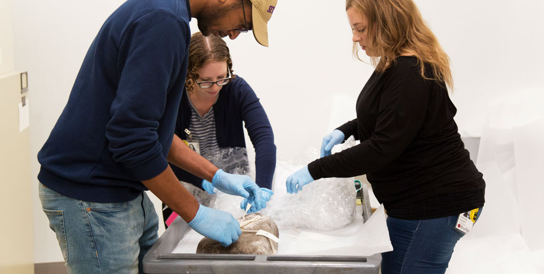 a man and two women wear gloves and carefully move a large stone object in the collection