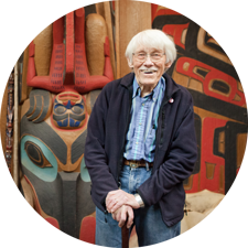 an older gentleman stands in front of his carvings inspired by Northwest Native Art