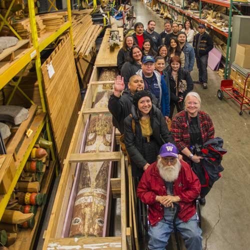 people stand around a carved wood canoe inside a storage building
