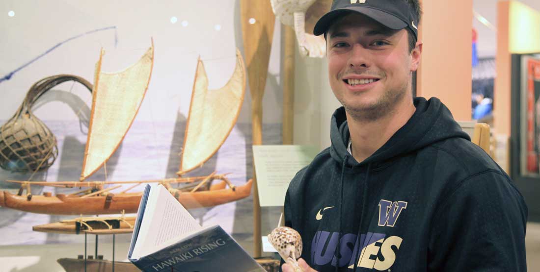 A young man wearing a husky hat and sweatshirt holds a book called Hawaiki Rising along with a museum object