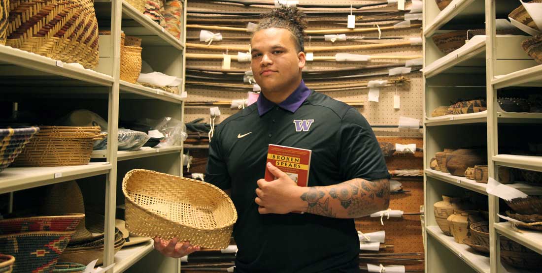 A young man stands in a row within the Native basket collection and holds a basket and book