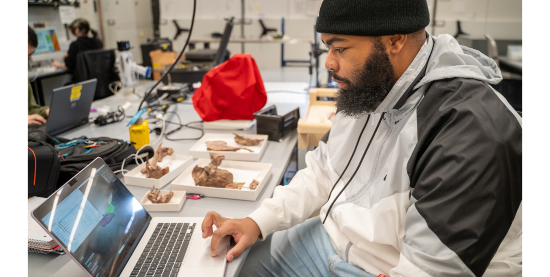 Man sits a table in front of his computer with several fossils sitting on the table.