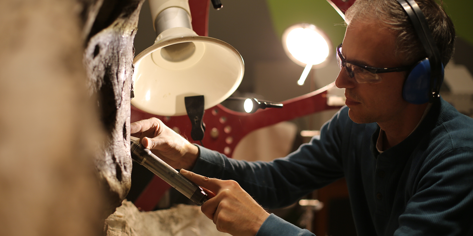 A fossil preparator wears safety goggles and ear protection while using a large air scribe to remove rock from the T. rex skull 