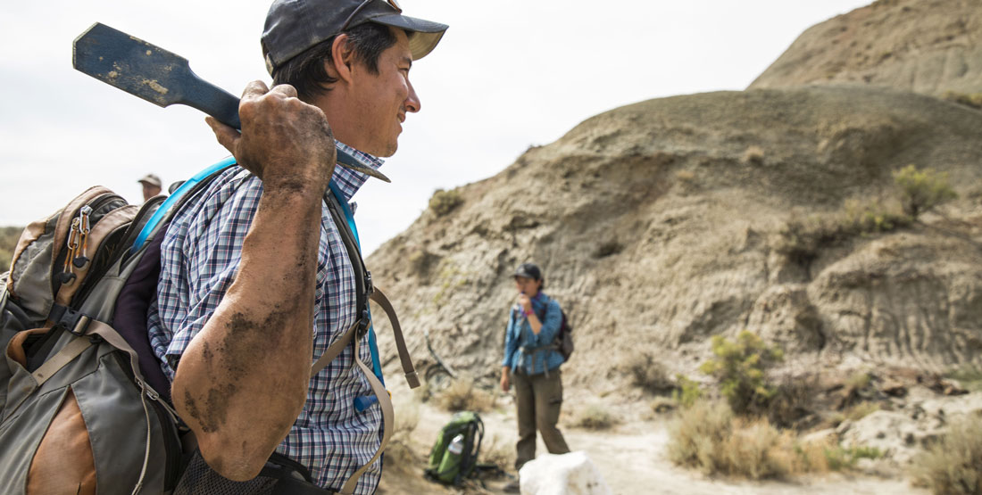 a man stands with a pickaxe over his shoulders and dirt covering his arms while doing fieldwork in the desert