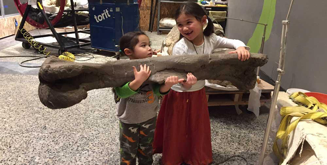 Two young girls hold a cast of a dinosaur bone that is double their size