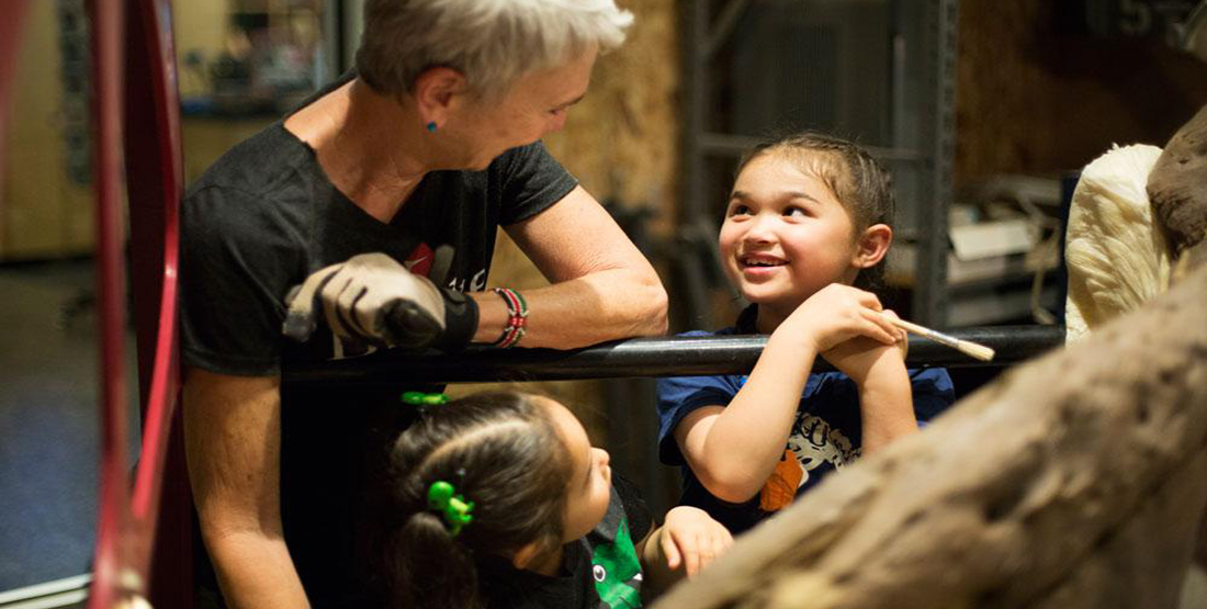 A young girl holding a paintbrush and a t.rex preparator smile at each other while a younger girl looks on
