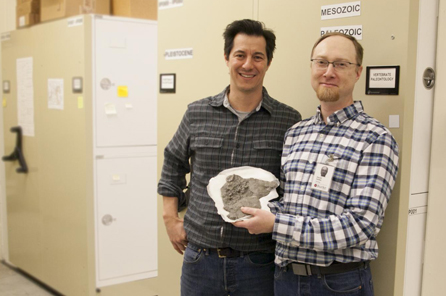 Two men stand amongst museum collections compactors and hold a small fossil