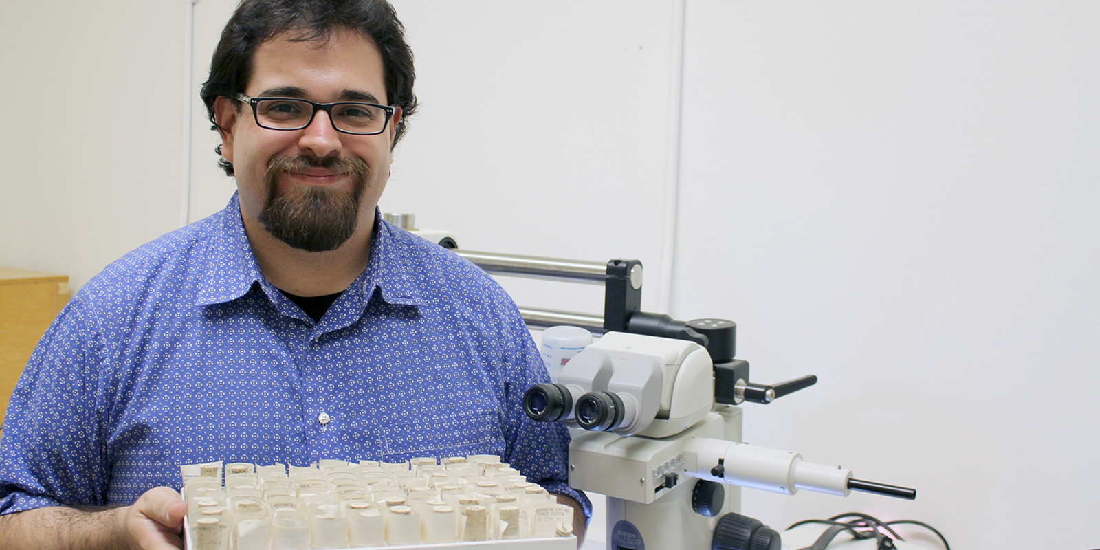 A male researcher holds a tray of tubes and stands next to a microscope