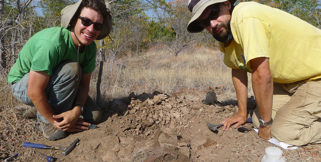 two men kneel next to a fossil in the dirt