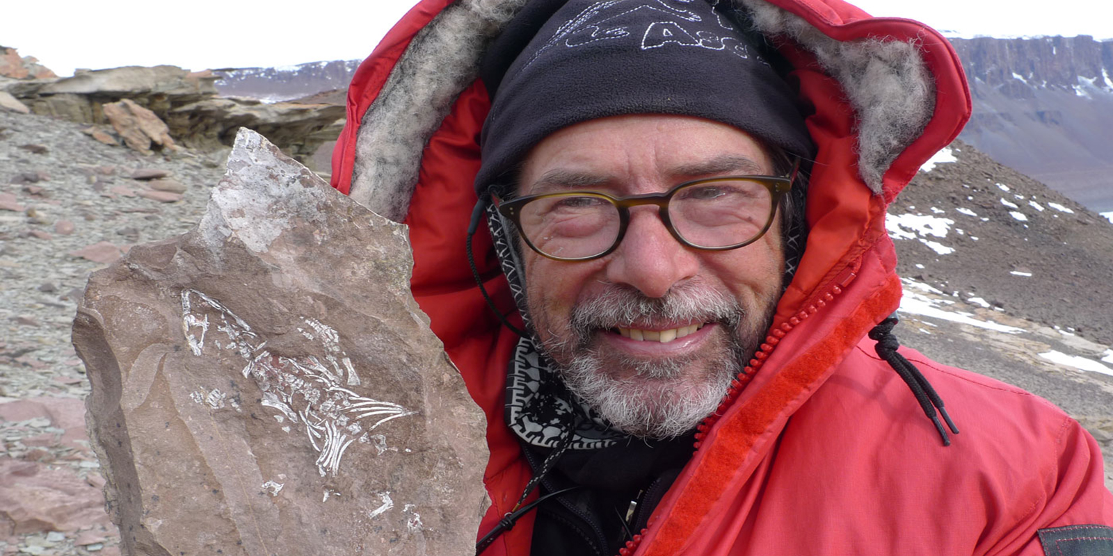 a man bundled in cold-weather gear holds up a fossil reptile visible in a slab of rock