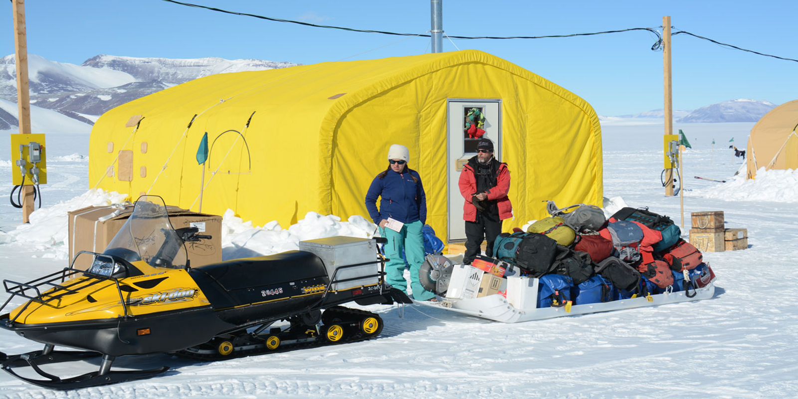 a woman and man stand with a snowmobile sled filled with gear