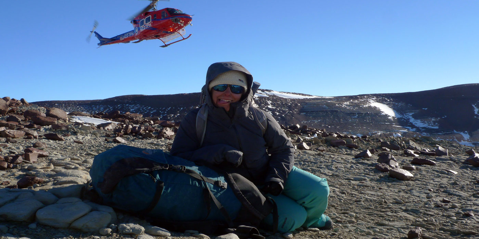 a woman bundled in cold-weather gear smiles at the camera as a helicopter lands behind her