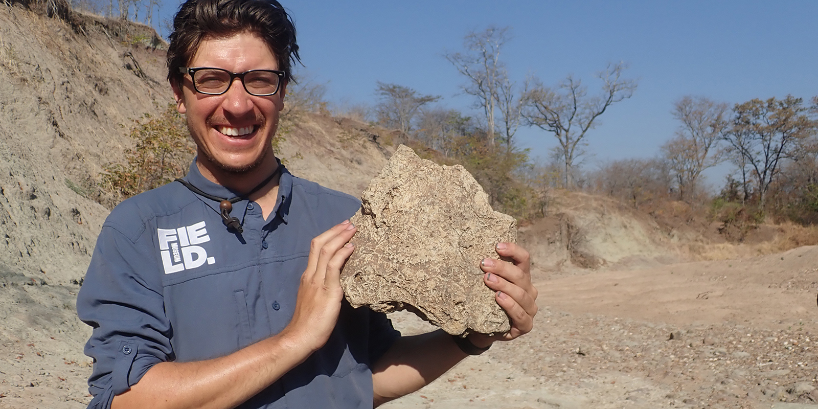 A young man smiles while holding a large fossil skull