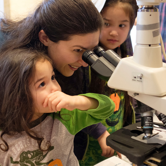 a mom looks into a microscope as her two girls look on