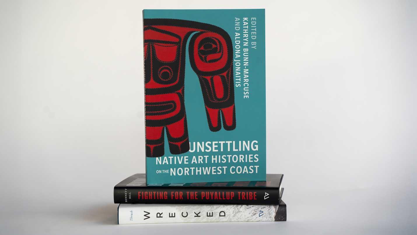 books stacked including Unsettling Native Art Histories on the Northwest Coast edited by Kathryn Bunn-Marcuse and Aldona Jonaitis, Fighting for the Puyallup Tribe: A Memoir by Ramona Bennett Bill, and Wrecked: Unsettling Histories from the Graveyard of the Pacific ​​​​​​​by Coll Thrush