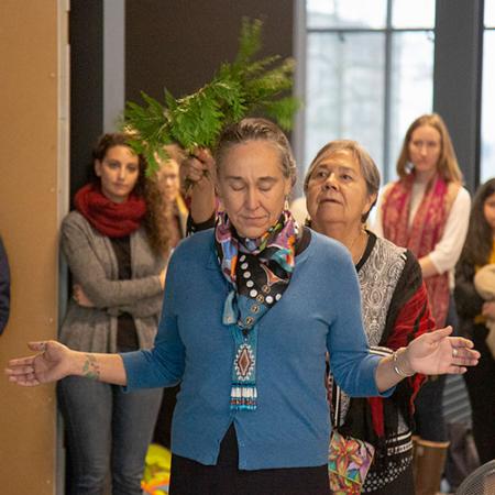 A woman stands with hands up and out as she is brushed with a cedar branch