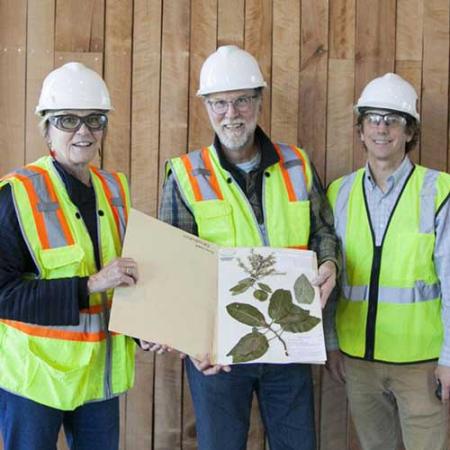 Three Burke Museum staff wear hardhats and safety vests while moving the first object into the new Burke Museum--a pressed plant specimen