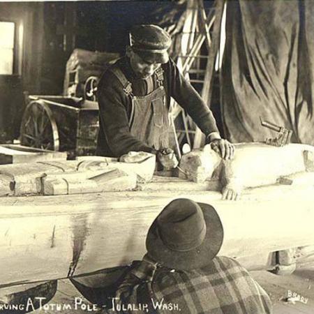 A old photograph of a man carving a totem pole in a workshop while another man looks on