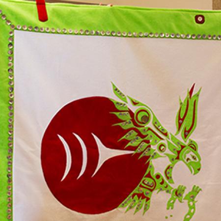 A button-blanket with green, white and red colors includes the Burke Museum logo and an eagle breaking out of chains