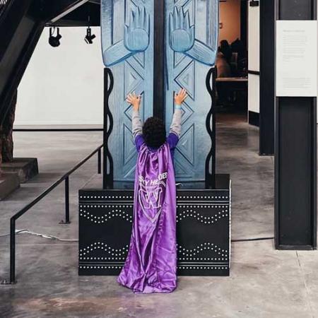 a child wearing a purple UW cape stands at the bottom of the weavers welcome sculpture in the burke museum lower lobby