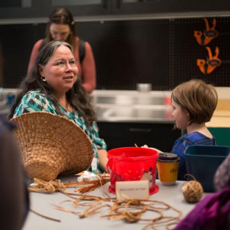 a woman speaks to a girl while weaving a cedar hat