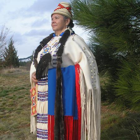 A woman wears full regalia and stands next to a tree