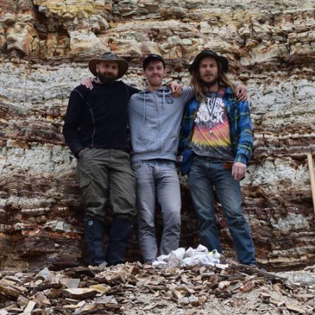 three young men stand in front of a rock wall showing many stratigraphic layers