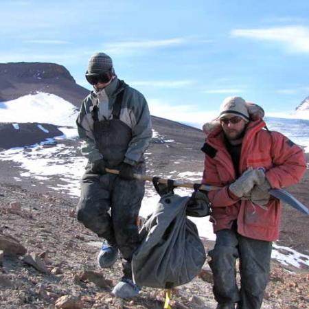 Two UW students use a shovel to carry a heavy fossil across barren landscape in Antarctica
