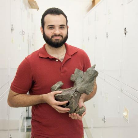 a young man stands within a row in the paleontology collections room and holds a fossil whale skull