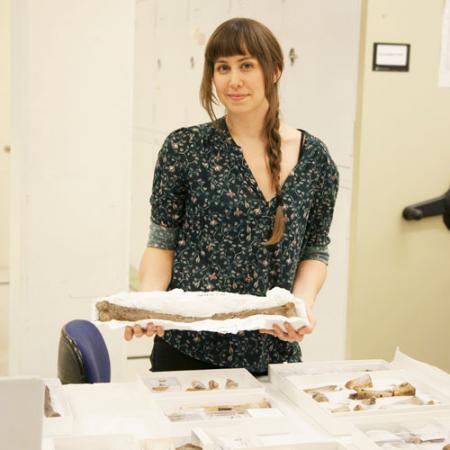 a young woman stands in front of a table full of fossils while holding a large fossil